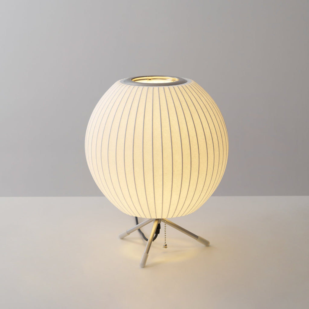 Round Clérachic table lamp lit on a grey set, warm light spilling under its shade and onto the white surface below.