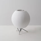An unlit round white Clérachic table lamp on a plain grey set, its pull chain hanging from the tripod base.