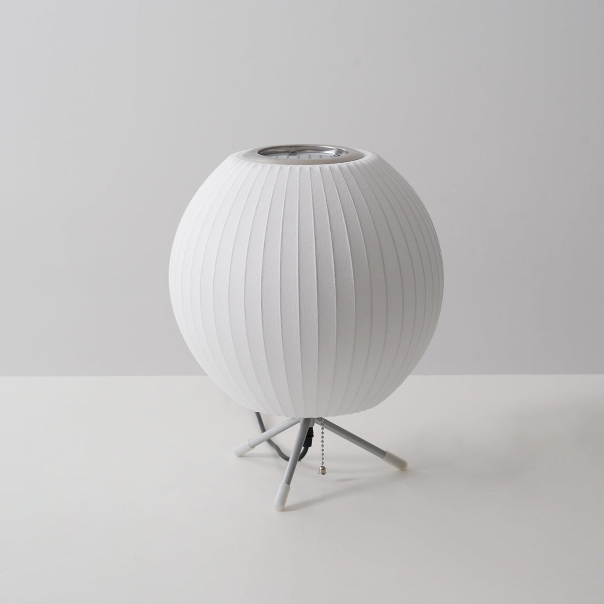 An unlit round white Clérachic table lamp on a plain grey set, its pull chain hanging from the tripod base.