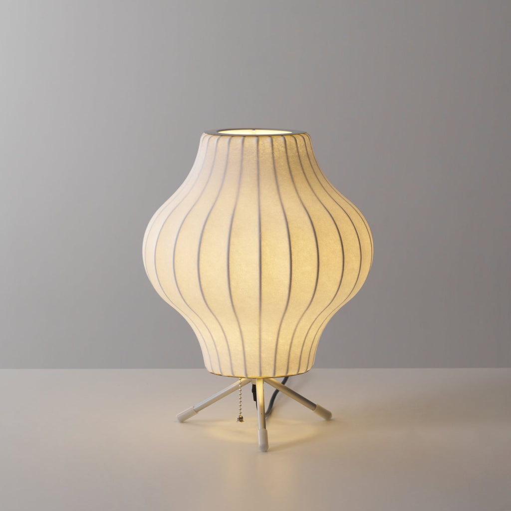 A pear-shaped Clérachic table lamp, lit, glowing warm on a pale grey set with light pooling around its tripod legs.