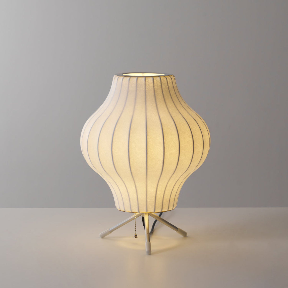 A pear-shaped Clérachic table lamp, lit, glowing warm on a pale grey set with light pooling around its tripod legs.