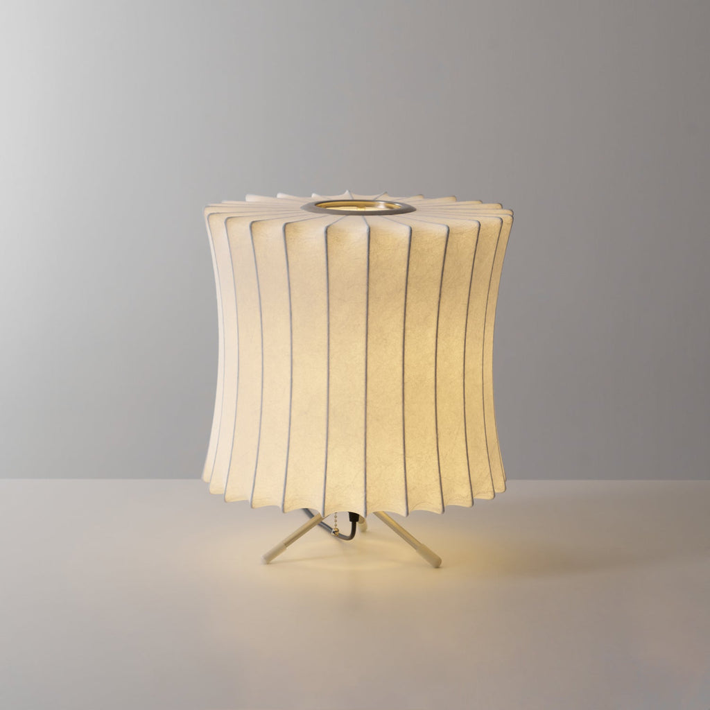 A lit waisted Clérachic table lamp with a pleated top edge, glowing warm against a grey set on a light surface.
