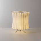A lit waisted Clérachic table lamp with a pleated top edge, glowing warm against a grey set on a light surface.