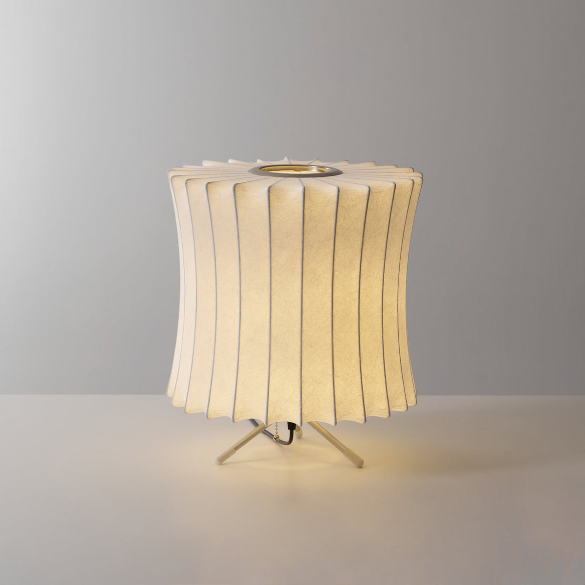 A lit waisted Clérachic table lamp with a pleated top edge, glowing warm against a grey set on a light surface.