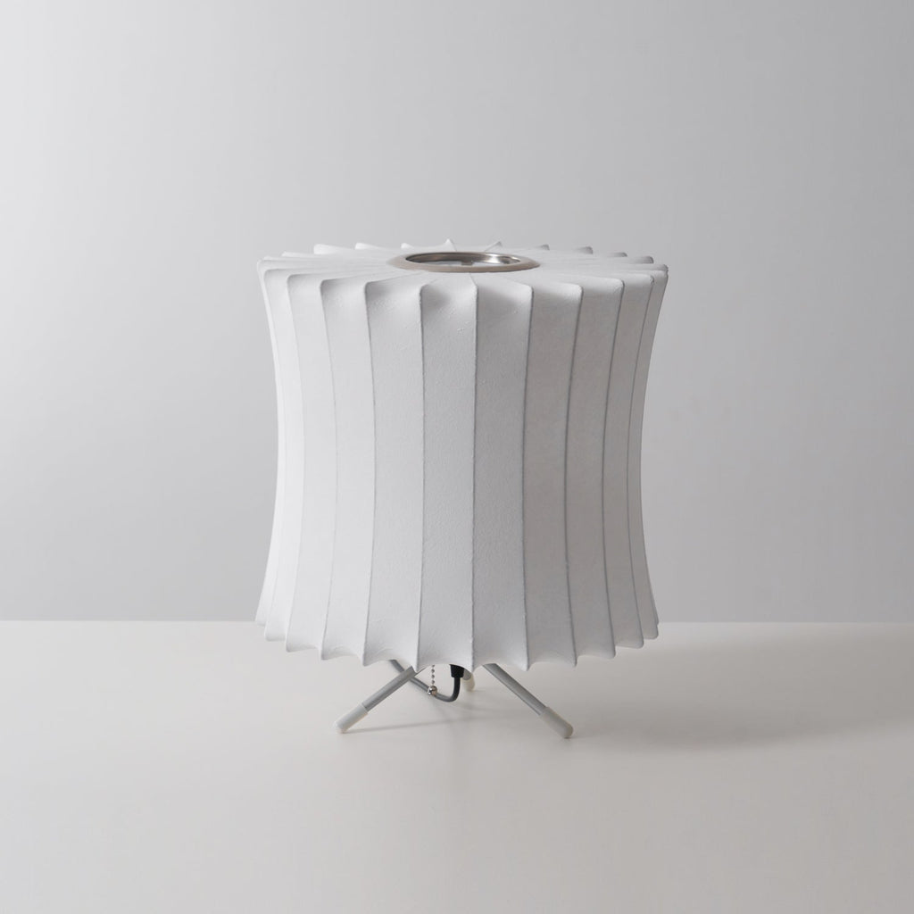 Unlit waisted white Clérachic table lamp with a pleated rim and a tripod stand, on a pale grey set.