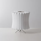 Unlit waisted white Clérachic table lamp with a pleated rim and a tripod stand, on a pale grey set.