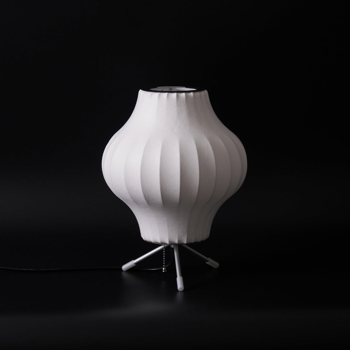 A pear-shaped white Clérachic table lamp, unlit, on a black set with a pull chain and a black cord at its base.