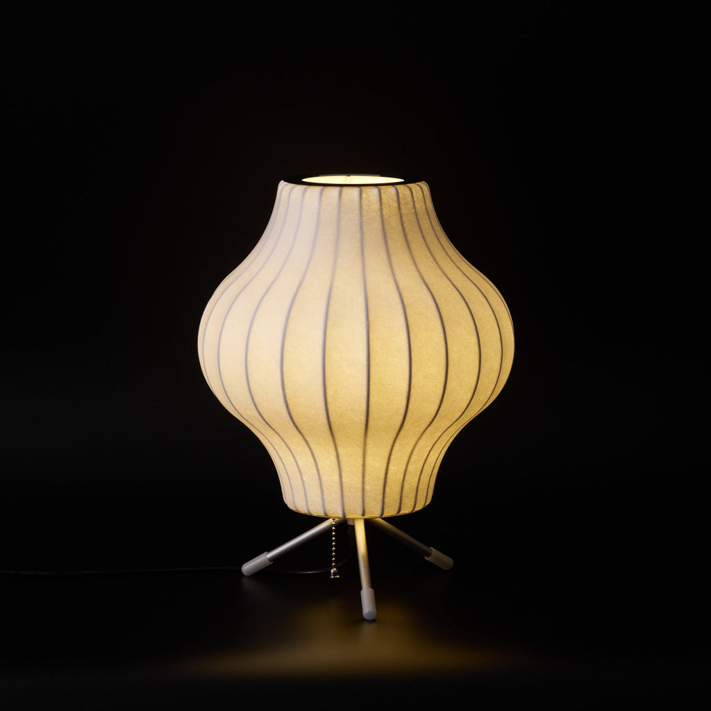 A glowing pear-shaped Clérachic table lamp on a black set, with warm light falling on the floor around its tripod.