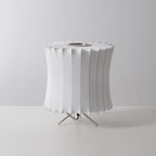 Waisted white Clérachic table lamp, unlit, seen from a slightly higher angle on a grey set, with a pleated top edge.