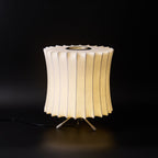 A waisted Clérachic table lamp lit warm against black, its pleated top edge and tripod legs sharply visible.