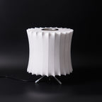 Unlit waisted white Clérachic table lamp on a black background, its pleated rim and ribbed shade clear to see.