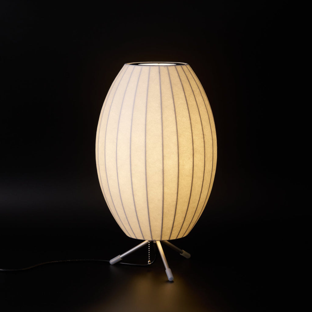 A glowing oval Clérachic table lamp against black, with its tripod legs, pull chain and black cord at the base.