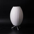 Unlit oval white Clérachic table lamp on a black set, its ribbed shade and white-tipped tripod legs in clear view.