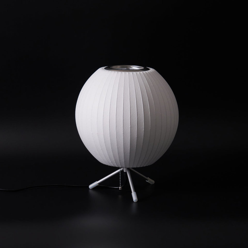 An unlit round white Clérachic table lamp on a black set, its tripod legs and a pull chain visible below.