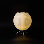 A round Clérachic table lamp, lit warm, against a black backdrop, with a glowing opening at the top of the shade.