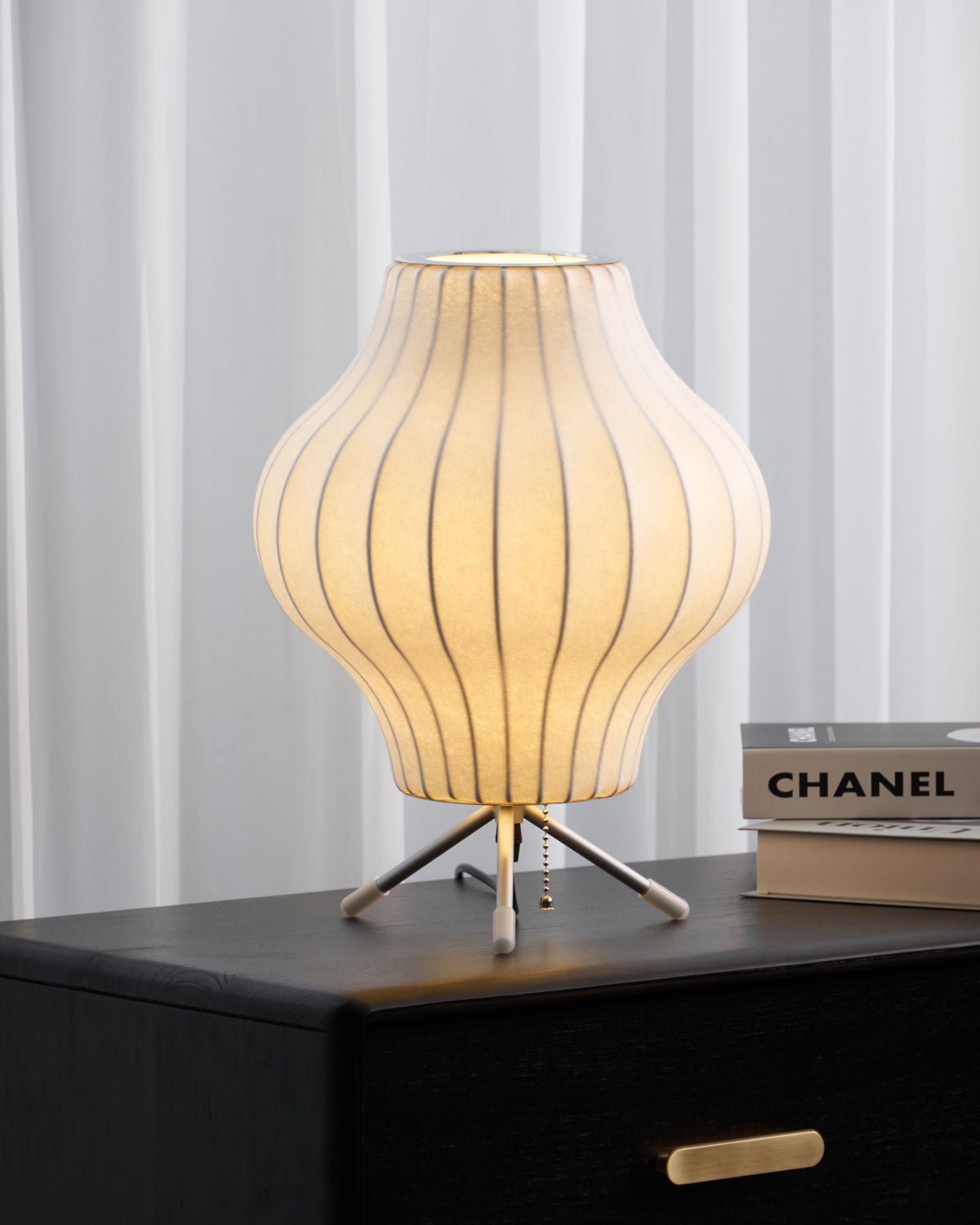 Pear-shaped Clérachic table lamp lit on a black drawer unit, a brass pull below and a stack of art books at the right.