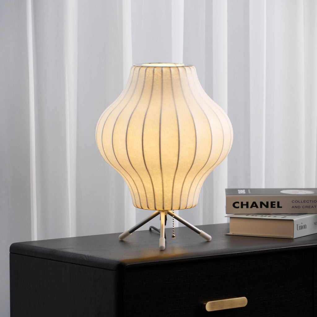 Lit pear-shaped Clérachic table lamp glowing warm on a black chest of drawers, with sheer curtains behind and art books beside it.