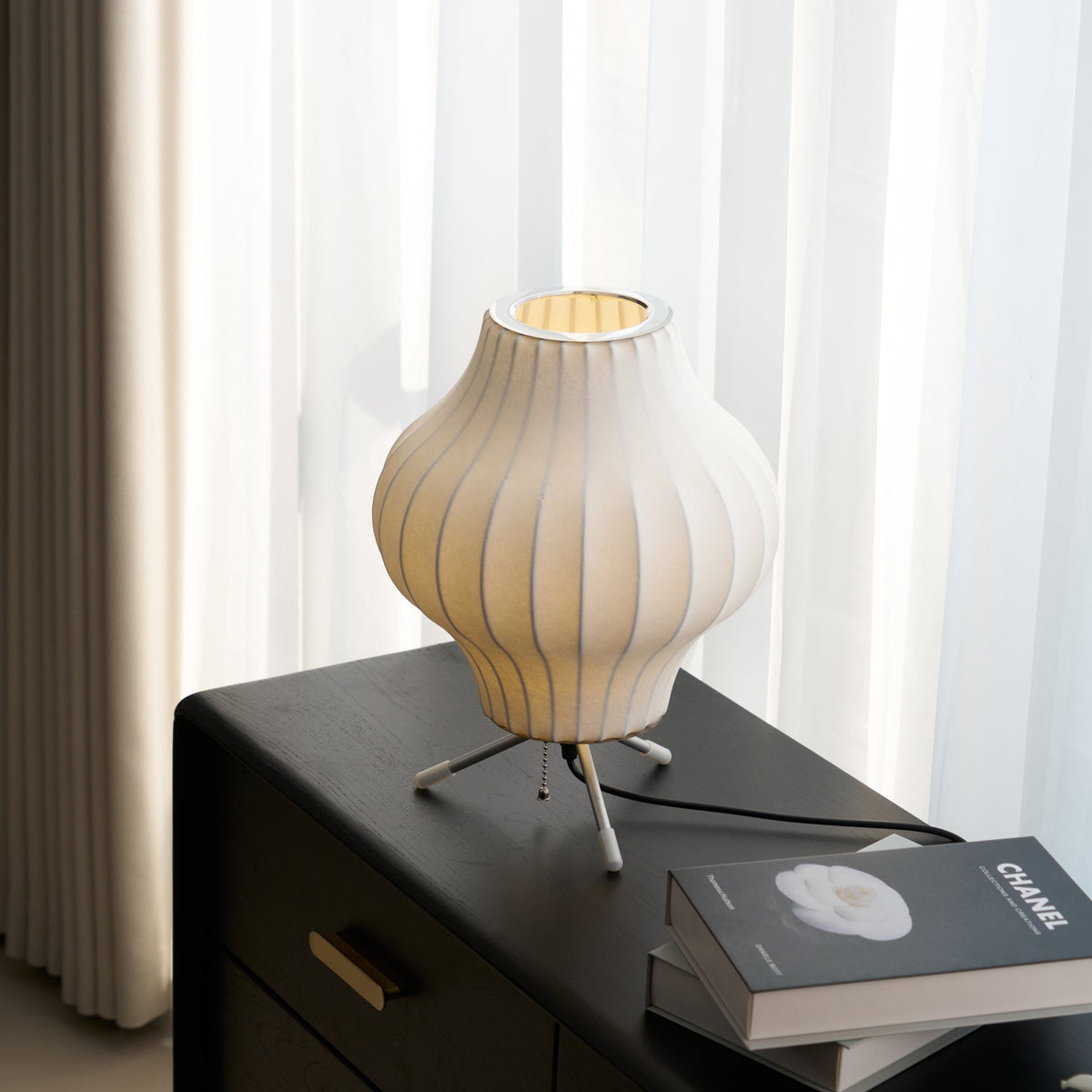 A white pear-shaped shade sits on a dark wooden chest by a curtained window, this Clérachic table lamp lit from within.