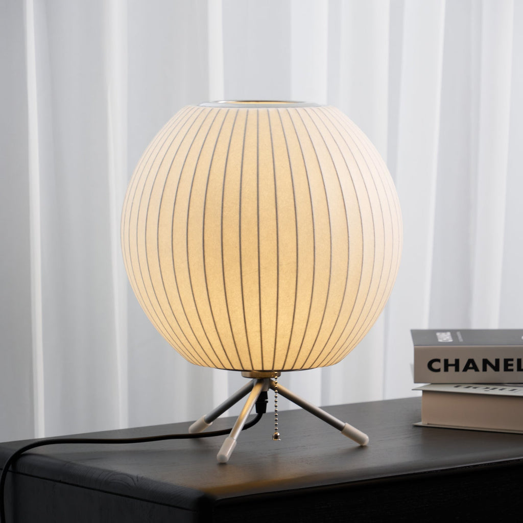 Round white ribbed shade on three slim legs, this Clérachic table lamp glows on a dark surface in front of sheer curtains.