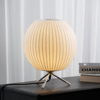 Round white ribbed shade on three slim legs, this Clérachic table lamp glows on a dark surface in front of sheer curtains.