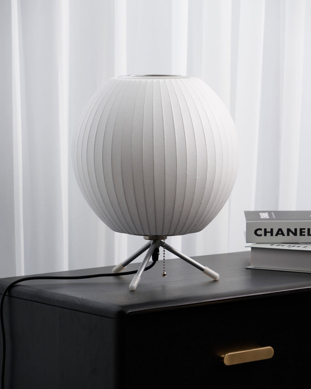 An unlit round white Clérachic table lamp on a black chest of drawers, with an art book beside it and white curtains behind.