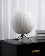 An unlit round white Clérachic table lamp on a black chest of drawers, with an art book beside it and white curtains behind.