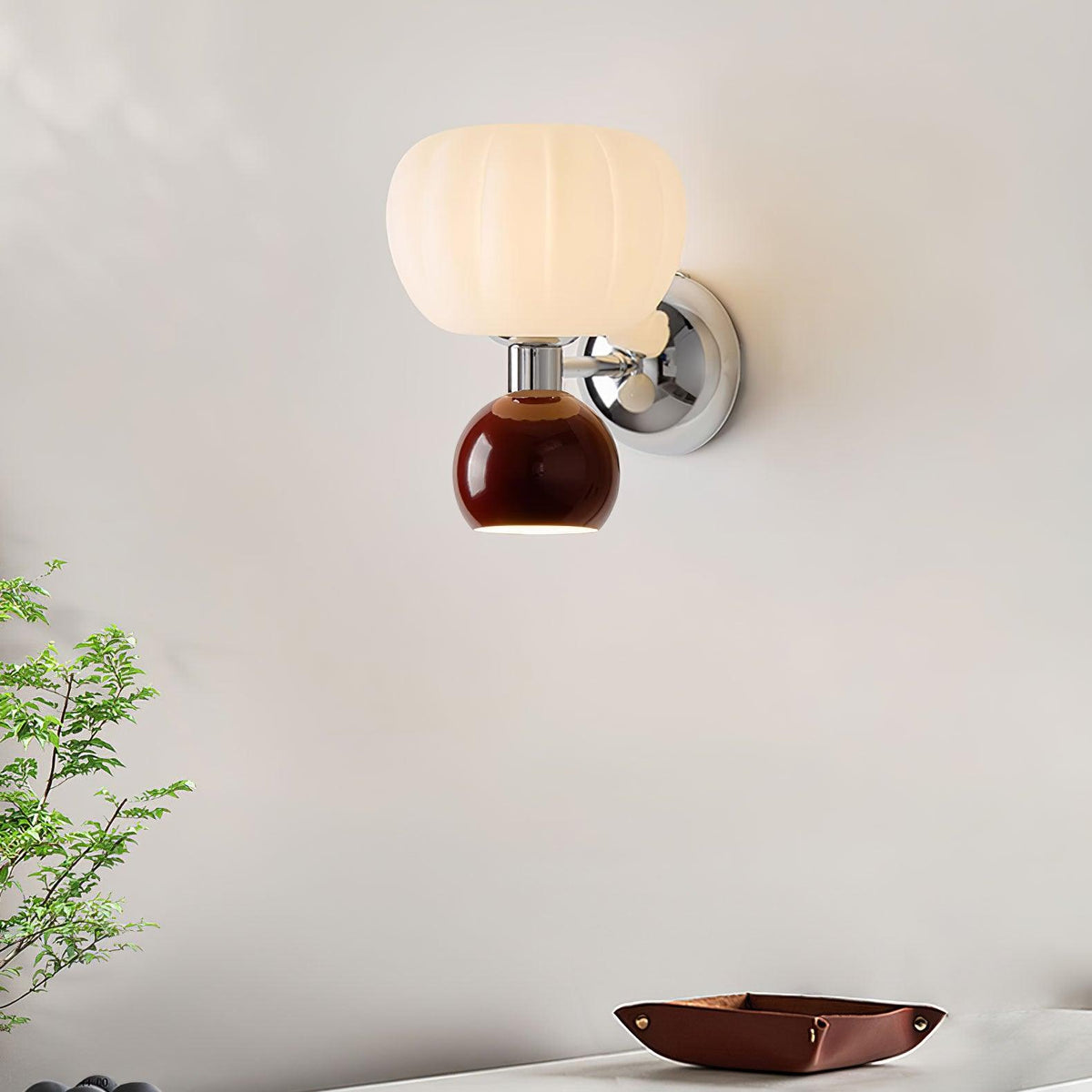 This Clérachic chrome wall sconce with a deep red ball glows on a white wall, with a green plant and a leather tray on a desk below.