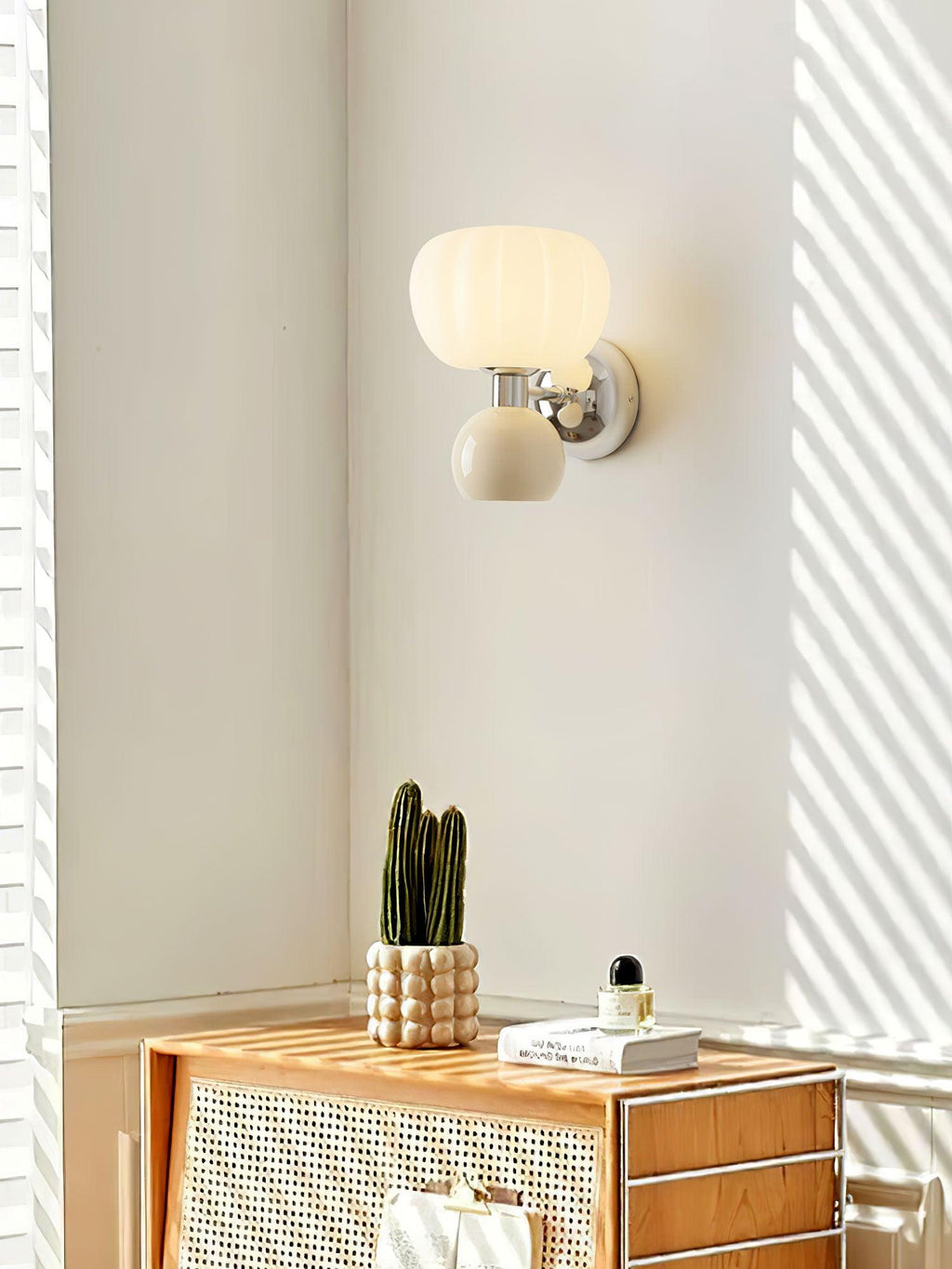 A Clérachic chrome wall light with a cream ball lit in a sunlit corner, with a cactus on a rattan-fronted cabinet below.