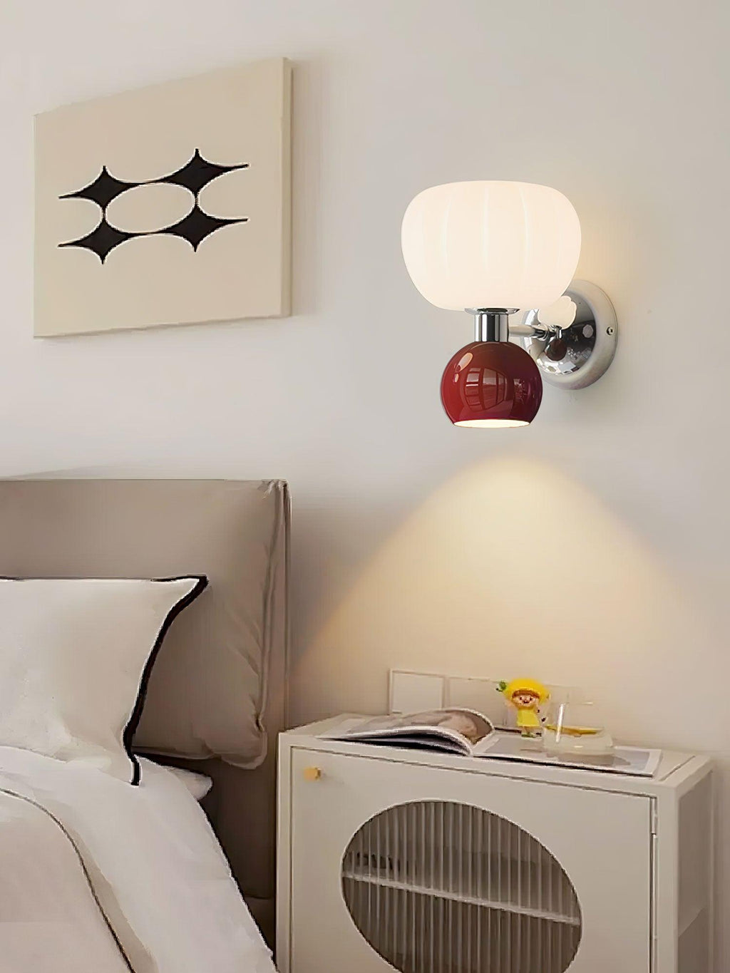 Above a cream bedside cabinet with a round cane panel, this Clérachic chrome wall light with a deep red ball throws a beam downward.