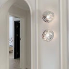 Two Clérachic wall lights with clear, melted-glass shades, stacked on a white wall beside an arched doorway.