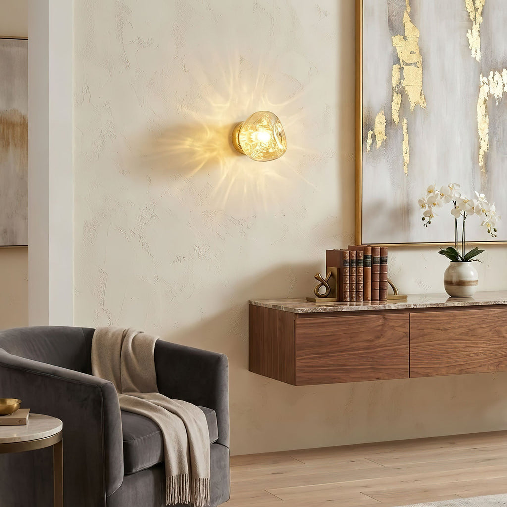 A small gold-toned Clérachic wall light glows above a floating walnut cabinet, beside a grey armchair and gold-leaf artwork.