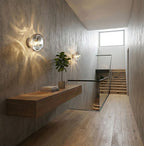 Along a concrete stairwell wall, a Clérachic wall light throws a pattern of light over a floating console and a potted tree.