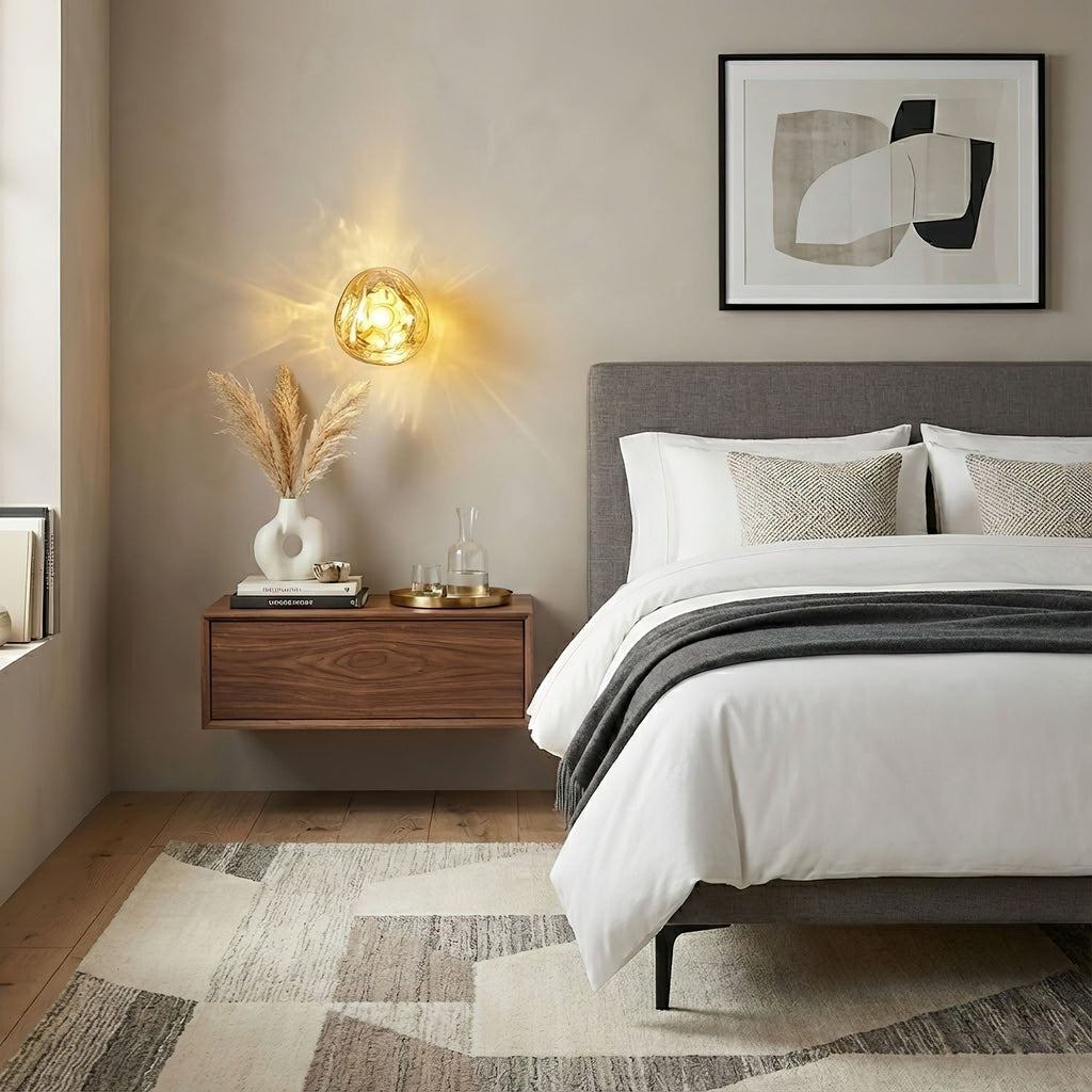 A gold-toned Clérachic wall light glows above a floating walnut bedside shelf, beside a bed with white and grey bedding.