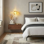 A gold-toned Clérachic wall light glows above a floating walnut bedside shelf, beside a bed with white and grey bedding.