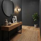 A Clérachic wall light with a clear glass shade sits on a dark wall above a walnut console, a round mirror and a jute rug.