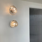 Two Clérachic wall lights, lit and stacked, with warm light showing through clear smoky melted-glass shades on a white wall.