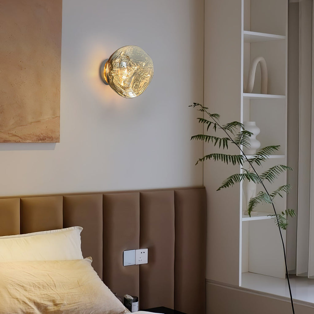 A Clérachic wall light with a clear melted-glass shade glows beside a tall plant and open shelves above a bed headboard.