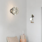 A Clérachic wall light with a clear, rippled-glass shade on a pale wall, with a small wall vase and a pastel cushion nearby.
