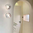 Two Clérachic wall lights with clear glass shades hang on a cream wall beside an arched doorway and a lit wall niche.