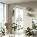 Two silver Clérachic wall lights with a reflective finish, on a ribbed beige wall beside an arch, with wooden chairs and plants.