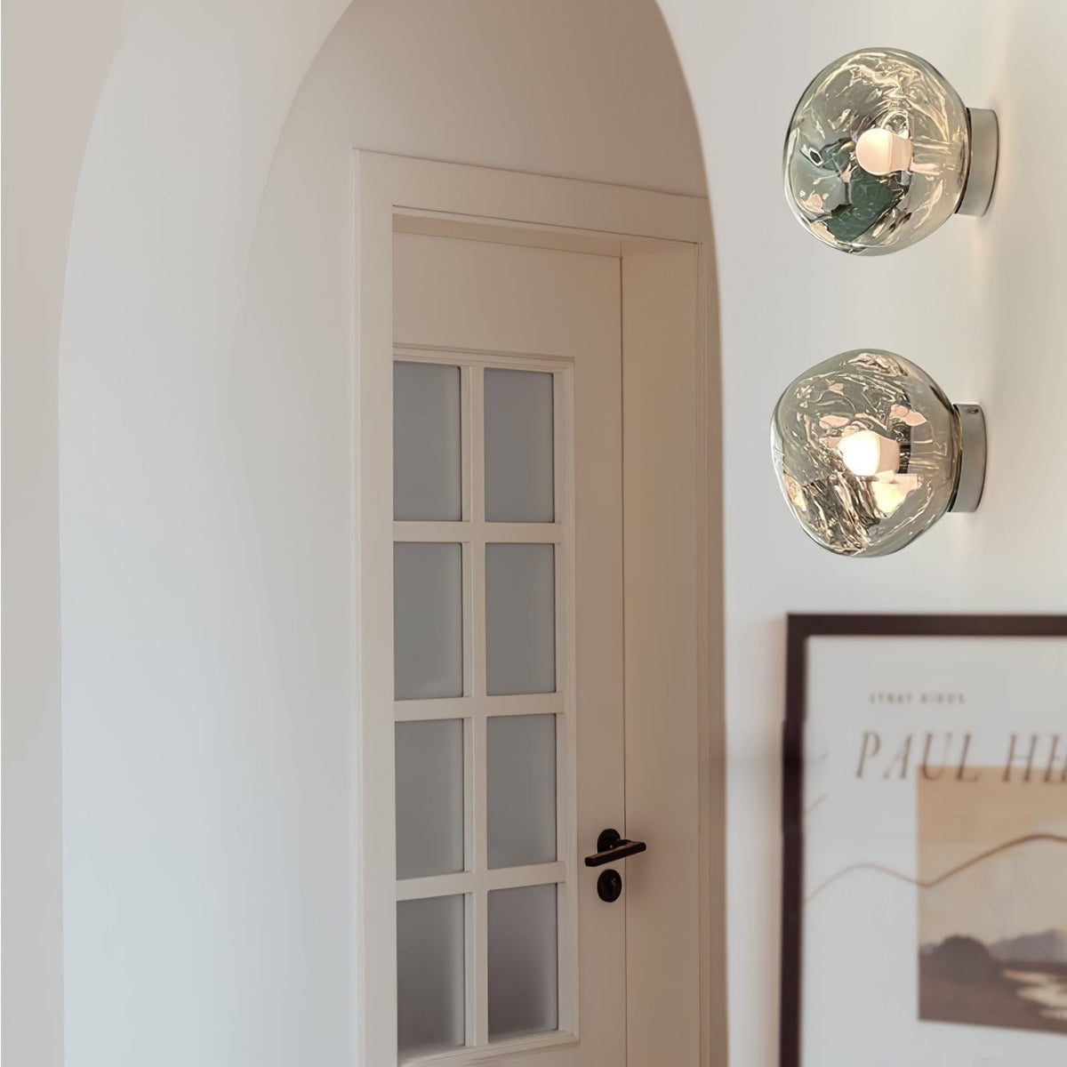 Two Clérachic wall lights with clear glass shades, stacked beside a panelled white door and a framed poster.