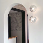 A pair of Clérachic wall lights with clear glass, mounted by a black-framed arched doorway with patterned wallpaper beyond.