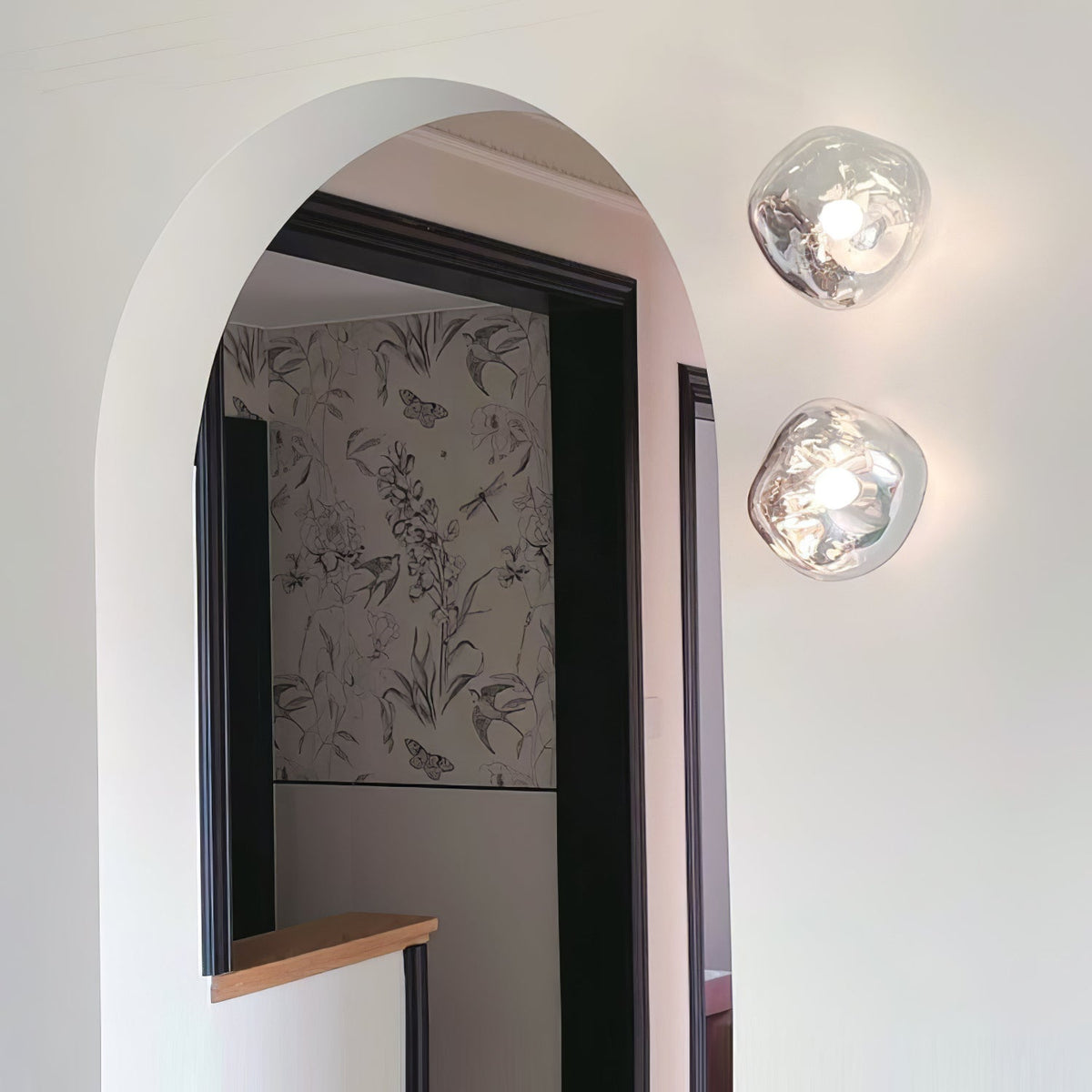 A pair of Clérachic wall lights with clear glass, mounted by a black-framed arched doorway with patterned wallpaper beyond.