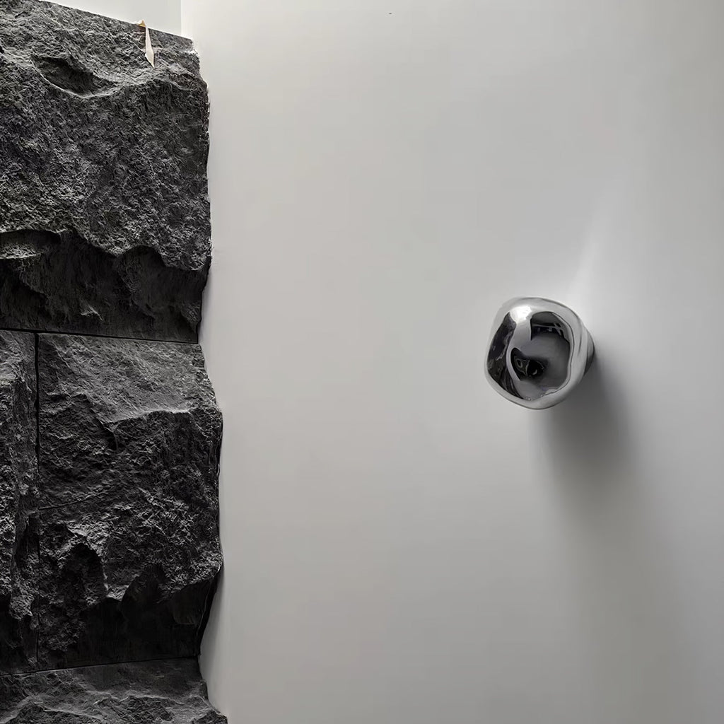 A silver, mirror-finish Clérachic wall light on a white wall, beside a rough dark stone surface.