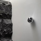 A silver, mirror-finish Clérachic wall light on a white wall, beside a rough dark stone surface.