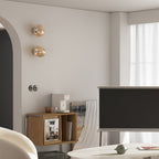 Two small amber-toned Clérachic wall lights, one above the other, in a living room with a low wooden unit and a TV.