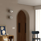 Two silver Clérachic wall lights stacked beside an arched wooden door, with a framed print and a black chair below.