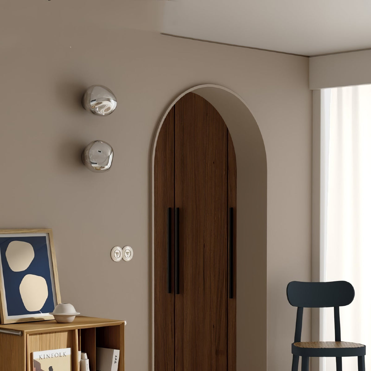 Two silver Clérachic wall lights stacked beside an arched wooden door, with a framed print and a black chair below.