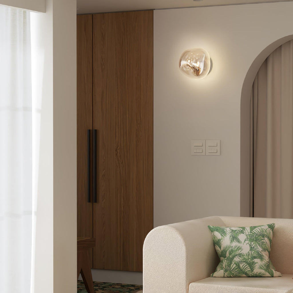 A lit Clérachic wall light with a champagne-toned glass shade, beside a wooden wardrobe and a cream armchair with a leaf cushion.
