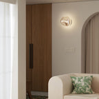 A lit Clérachic wall light with a champagne-toned glass shade, beside a wooden wardrobe and a cream armchair with a leaf cushion.
